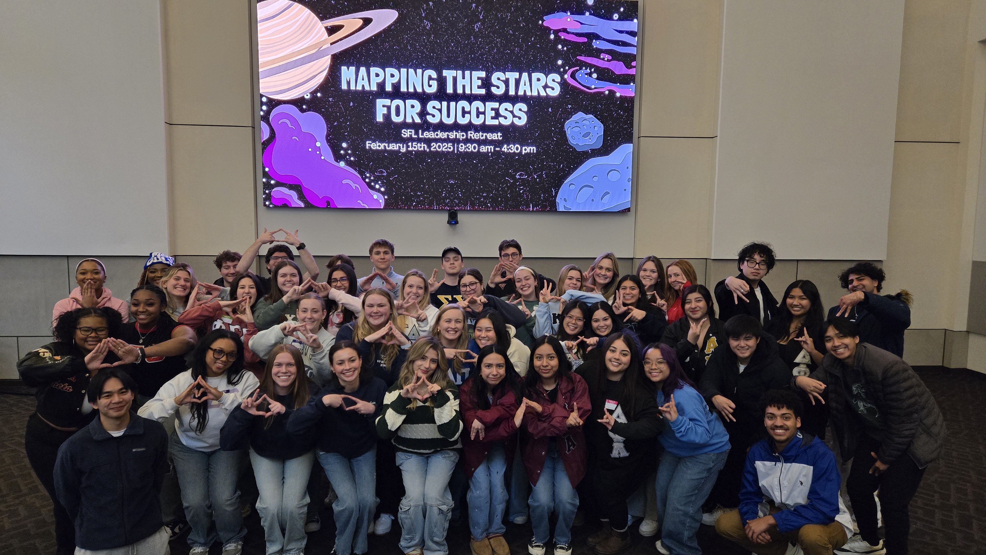 Students gathered together posing for a picture in a large room in front of a large flat screen
